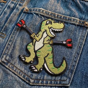 May include: A green and white embroidered dinosaur patch with a smiling face and holding two black and red tools, sewn onto a blue denim pocket.