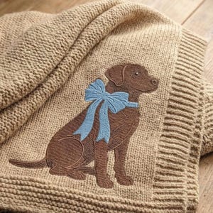 May include: A tan knitted blanket featuring a brown Labrador Retriever with a light blue bow. The dog is embroidered on the blanket, which has a ribbed border and a soft, textured appearance. The blanket is folded, showcasing its cozy design.