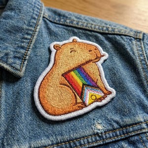 May include: Embroidered patch featuring a capybara holding a rainbow Progress Pride flag. The patch has a white border and is sewn onto a denim jacket. The capybara is brown with a white outline.