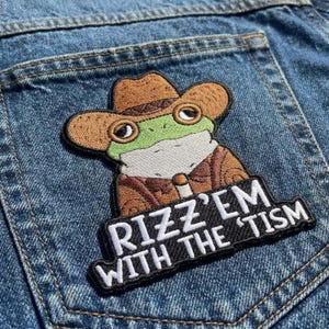 May include: Embroidered patch featuring a cartoon frog wearing a cowboy hat and jacket. The frog is green and brown, with the text "RIZZ'EM WITH THE 'TISM" in white on a black background. The patch is on a denim pocket.