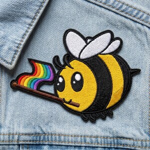 May include: Embroidered patch featuring a cartoon bee with a rainbow flag. The bee has a yellow body with black stripes, white wings, and large, friendly eyes. The patch is on a light blue denim background.