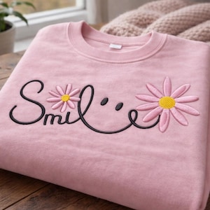 Könnte beinhalten: Hellrosa Sweatshirt mit dem Wort "Smile" in schwarzer Schrift, verziert mit zwei gestickten Gänseblümchen in Rosa und Gelb. Das Sweatshirt liegt gefaltet auf einer Holzoberfläche.