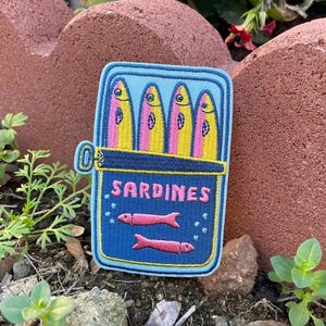 May include: A blue embroidered patch featuring a sardine can design. The can is open, revealing four colorful sardines with pink, yellow, and blue details. The word "SARDINES" is printed in pink on the can. Two pink fish are below the sardines.