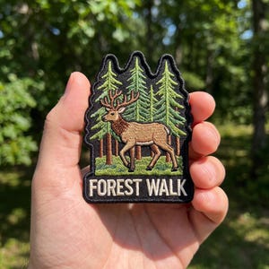 May include: An embroidered patch featuring a tan elk with large antlers standing in a green forest of pine trees. The patch has a black border and the text FOREST WALK in white capital letters at the bottom against a black background.