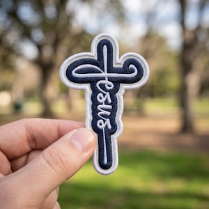 May include: A navy blue and white embroidered patch in the shape of a cross. The word "Jesus" is written in cursive script down the center of the cross. The patch is held in a person's hand against a blurred outdoor background.