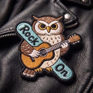 May include: An embroidered patch featuring a brown and white owl playing a tan acoustic guitar. The owl is positioned over a light blue banner with the black text "Rock On" written across it. The patch has a black border and detailed stitching.