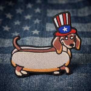 May include: Embroidered patch featuring a dachshund in a hot dog bun, wearing a patriotic Uncle Sam hat. The hat is red, white, and blue with a star. The dog has brown and tan fur, with a black outline.