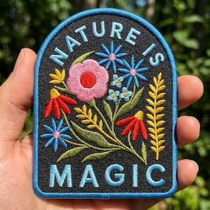 May include: An embroidered patch with a black background and a blue border. The text NATURE IS MAGIC is stitched in light blue. The design features a central pink flower, blue and red wildflowers, and yellow foliage with green stems and leaves.