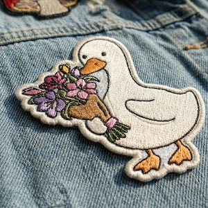 May include: Embroidered patch featuring a white goose holding a bouquet of flowers. The goose has an orange beak and feet, with a detailed outline. The flowers are in shades of pink, purple, and yellow. The patch is on a denim background.