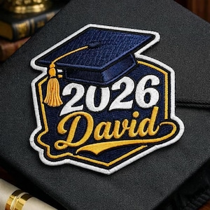 May include: Embroidered patch featuring a graduation cap design with the text "2026 David." The patch has a navy blue cap with a gold tassel, white and gold lettering, and a navy blue and gold border. The patch is on a black surface.