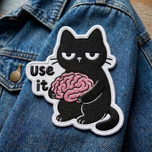 May include: Embroidered patch featuring a black cat holding a pink brain. The cat has a grumpy expression and white accents. The patch includes the text "Use it" in a white bubble. The patch is on a blue denim jacket.
