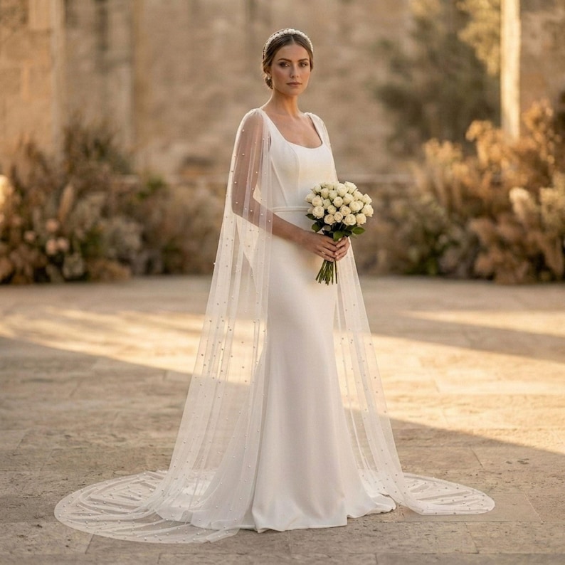 Peut inclure: Une robe de mari&eacute;e blanche avec une longue tra&icirc;ne et une cape transparente orn&eacute;e de petites perles. La robe a un design simple et &eacute;l&eacute;gant avec un corsage ajust&eacute; et une jupe fluide. Un bouquet de fleurs blanches est tenu.