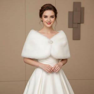 May include: A white faux fur shawl with a decorative rhinestone clasp. The shawl is draped over a white dress, creating an elegant look. The image features a woman wearing the shawl, with a neutral background.