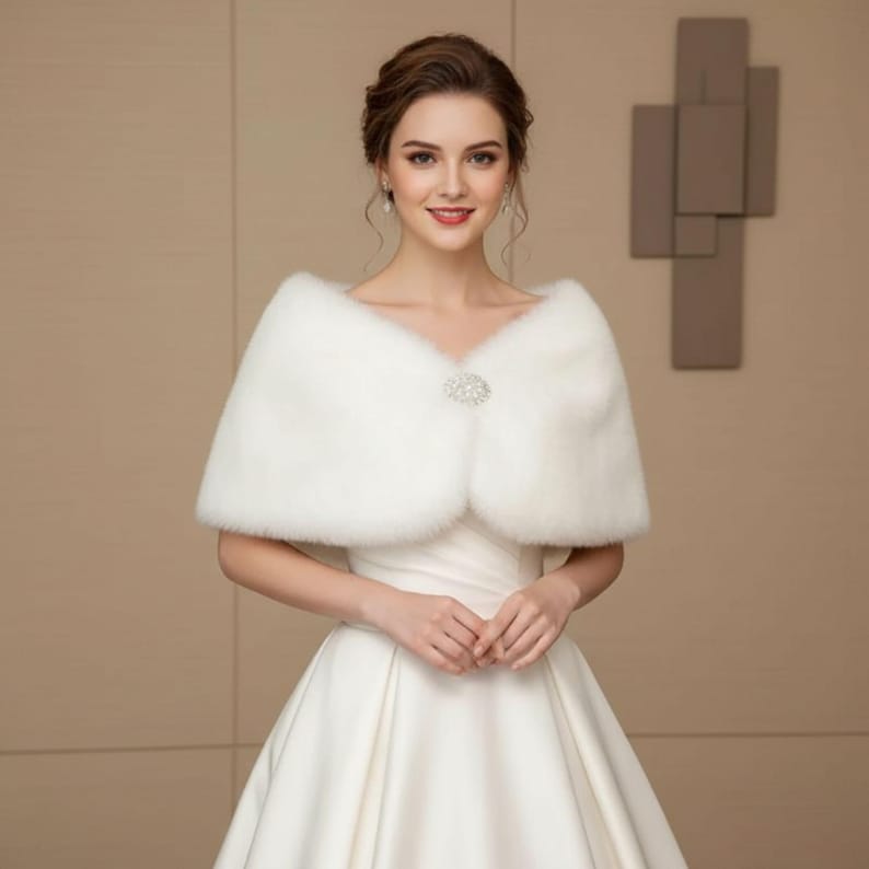 May include: A white faux fur shawl with a decorative rhinestone clasp. The shawl is draped over a white dress, creating an elegant look. The image features a woman wearing the shawl, with a neutral background.