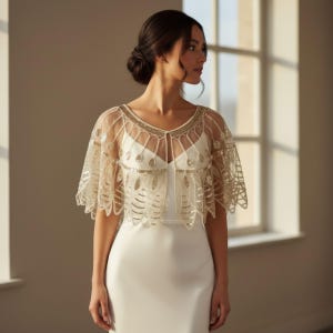 May include: A sheer, ivory-colored capelet with intricate gold beading and embellishments. The capelet features a delicate, open design with a scalloped edge, perfect for bridal or formal wear. The capelet is worn over a white dress.