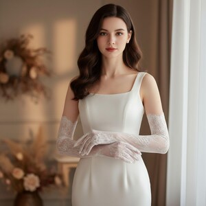 May include: A white, sleeveless wedding dress with a square neckline. The bride is wearing long, white lace gloves. The background is softly lit, with a window and floral arrangements visible.