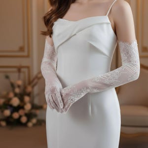 May include: A white, sleeveless dress with a draped bodice and spaghetti straps. The dress is paired with long, sheer lace gloves. The gloves feature intricate floral lace patterns, adding a delicate touch to the ensemble. The overall style is elegant and suitable for formal occasions.