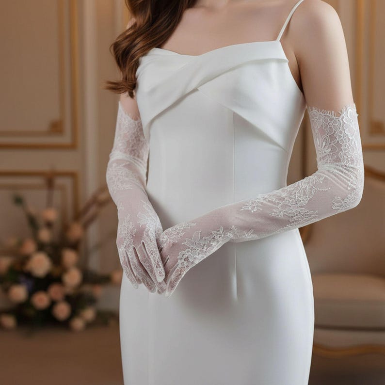 May include: A white, sleeveless dress with a draped bodice and spaghetti straps. The dress is paired with long, sheer lace gloves. The gloves feature intricate floral lace patterns, adding a delicate touch to the ensemble. The overall style is elegant and suitable for formal occasions.