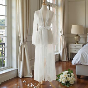 Op de afbeelding: Een witte, doorschijnende kanten robe en bijpassende lange rok worden op een mannequin getoond. De robe heeft lange mouwen en een strik in de taille. Een boeket witte rozen ligt op de vloer. De setting is een lichte kamer met grote ramen en crèmekleurige gordijnen.