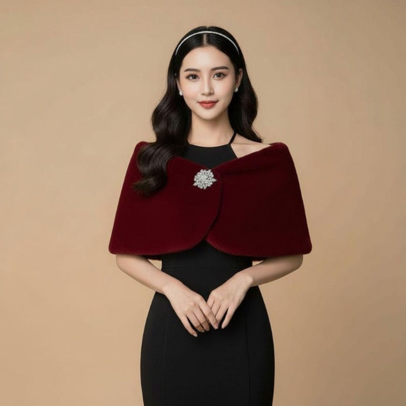 May include: A burgundy velvet cape with a decorative brooch. The cape is draped over the shoulders and fastened with a sparkling, silver-toned brooch. The woman is wearing a black dress and a silver headband.