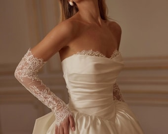 May include: An ivory strapless gown featuring a ruched bodice with a sweetheart neckline adorned with white pearls. The dress is paired with long, sheer white lace gloves that extend to the mid-forearm, showcasing intricate floral patterns.