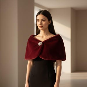 May include: A burgundy faux fur capelet with a decorative rhinestone brooch. The capelet is draped over a black dress. The image is well-lit, showcasing the texture and color of the capelet.