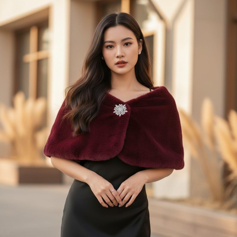 May include: A burgundy faux fur capelet with a sparkling rhinestone brooch. The capelet is draped over the shoulders and fastened at the front. The garment is a stylish accessory for formal events.