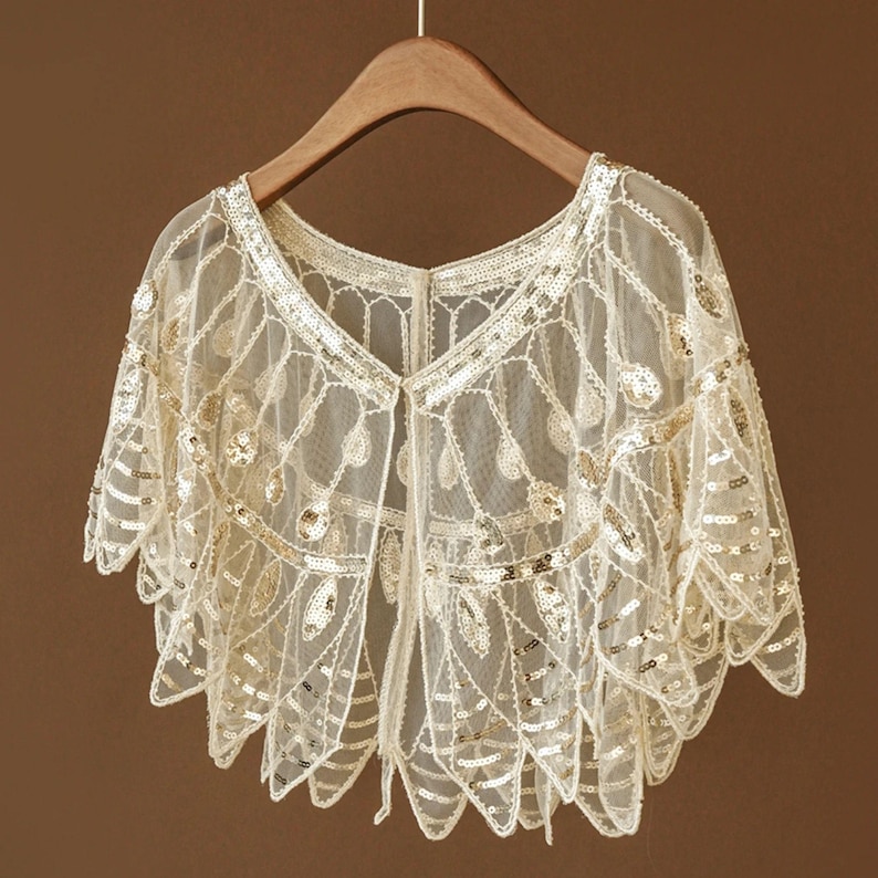 May include: A cream-colored, open-front bolero with intricate lace detailing and gold sequins. The bolero is hanging on a wooden hanger against a brown background. The delicate lace pattern and shimmering sequins create an elegant, vintage aesthetic.