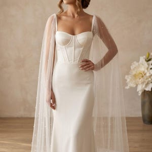 Peut inclure: Une robe de mari&eacute;e blanche avec un corsage ajust&eacute; et une jupe fluide. La robe est orn&eacute;e d'une cape transparente parsem&eacute;e de petites perles. La robe a un d&eacute;collet&eacute; en c&oelig;ur et un corsage de style corset. Le design est &eacute;l&eacute;gant et sophistiqu&eacute;.