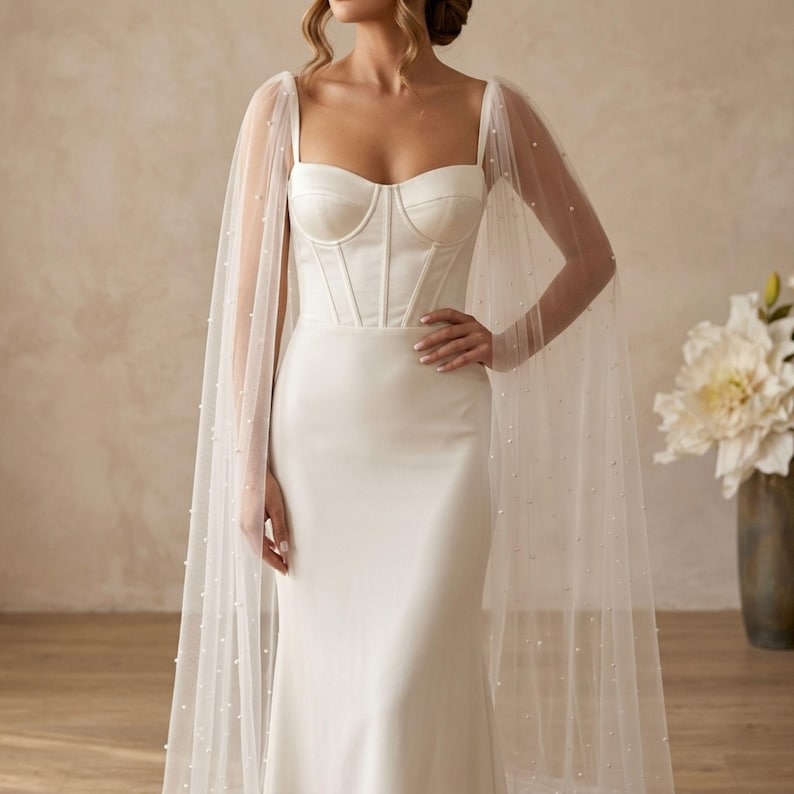 Peut inclure: Une robe de mari&eacute;e blanche avec un corsage ajust&eacute; et une jupe fluide. La robe est orn&eacute;e d'une cape transparente parsem&eacute;e de petites perles. La robe a un d&eacute;collet&eacute; en c&oelig;ur et un corsage de style corset. Le design est &eacute;l&eacute;gant et sophistiqu&eacute;.