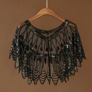 May include: Black sequined cape with an intricate, Art Deco-inspired design. The sheer fabric is embellished with sequins and hangs from a wooden hanger. The cape features a scalloped hemline and a delicate, open weave.