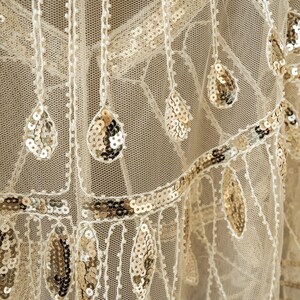 May include: Close-up of a cream-colored fabric with intricate detailing. The fabric features a delicate net-like structure adorned with shimmering gold sequins in teardrop and linear patterns. The design evokes a vintage or art deco aesthetic.