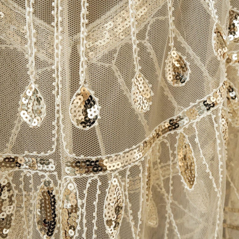 May include: Close-up of a cream-colored fabric with intricate detailing. The fabric features a delicate net-like structure adorned with shimmering gold sequins in teardrop and linear patterns. The design evokes a vintage or art deco aesthetic.