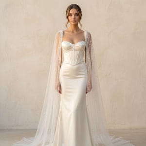 Peut inclure: Une robe de mari&eacute;e en satin blanc avec un d&eacute;collet&eacute; en c&oelig;ur et un corsage ajust&eacute;. La robe pr&eacute;sente une longue jupe fluide et est associ&eacute;e &agrave; une cape transparente orn&eacute;e de petites perles &eacute;parpill&eacute;es. La mari&eacute;e se tient devant un fond neutre.