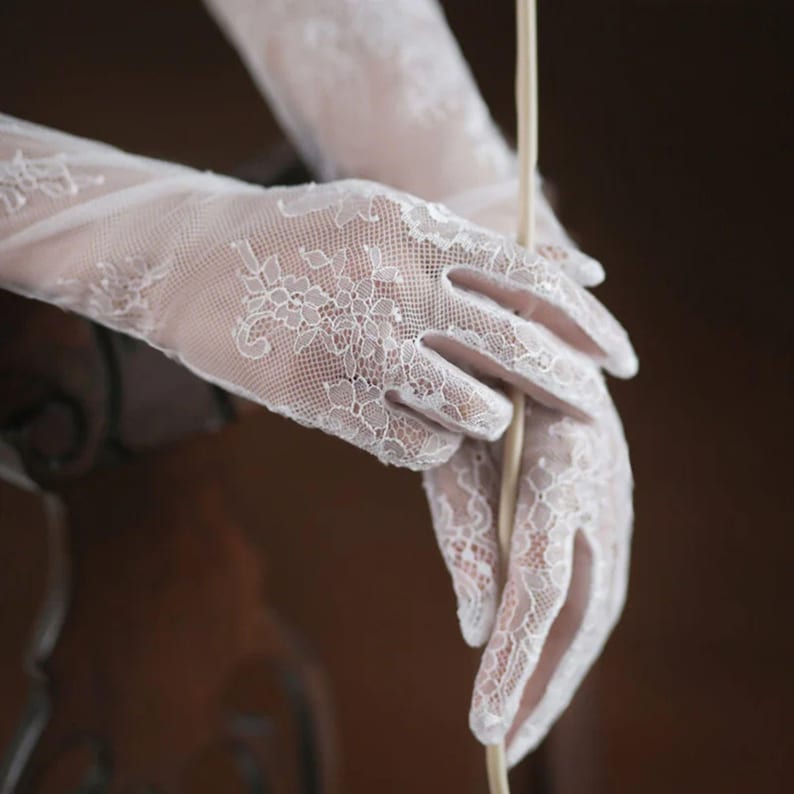 May include: Elegant white lace gloves with floral patterns. The gloves are long, extending past the wrists, and feature intricate detailing. The delicate fabric and design suggest a formal or bridal aesthetic.