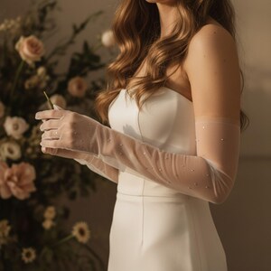 Puede incluir: Un vestido de novia blanco sin tirantes con guantes largos y transparentes adornados con pequeños detalles brillantes. Los guantes, salpicados de pedrería, se extienden más allá de los codos. Arreglos florales suaves y borrosos en el fondo.