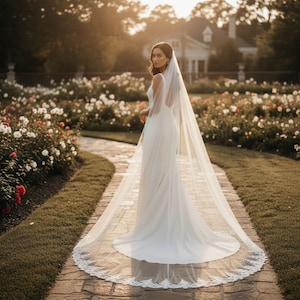 May include: A woman in a white wedding dress and veil stands on a brick path in a garden. The dress is sleeveless with a flowing skirt. The veil is long and edged with lace. The garden features rose bushes and a house in the background.