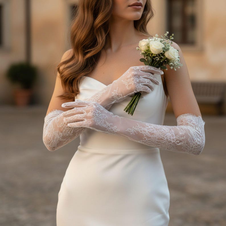 May include: Elegant white lace gloves with floral patterns, extending to the upper arm. The gloves are paired with a strapless white dress and a small bouquet of white roses and greenery. The overall aesthetic is bridal and romantic.