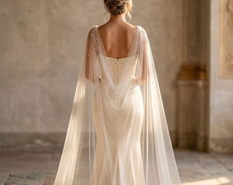 May include: A cream-colored wedding dress with a sheer, pearl-studded cape. The dress has a fitted bodice and a flowing skirt. The cape drapes over the shoulders and down the back, creating an elegant and romantic look. The dress is worn by a person with their back to the camera.