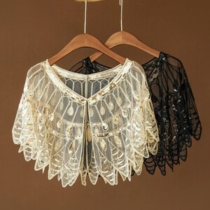 May include: Two sequined shawls on wooden hangers. One is a cream color with gold sequins, and the other is black. Both have a delicate, lacy design with a scalloped edge. The shawls are displayed against a brown background.