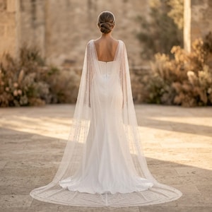 Peut inclure: Robe de mari&eacute;e blanche avec une longue tra&icirc;ne et une cape transparente orn&eacute;e de petites perles. La robe a un d&eacute;collet&eacute; carr&eacute; et une silhouette ajust&eacute;e. La cape flotte gracieusement, cr&eacute;ant un look &eacute;l&eacute;gant et romantique.