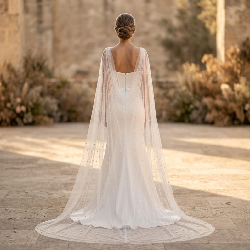 Peut inclure: Robe de mari&eacute;e blanche avec une longue tra&icirc;ne et une cape transparente orn&eacute;e de petites perles. La robe a un d&eacute;collet&eacute; carr&eacute; et une silhouette ajust&eacute;e. La cape flotte gracieusement, cr&eacute;ant un look &eacute;l&eacute;gant et romantique.