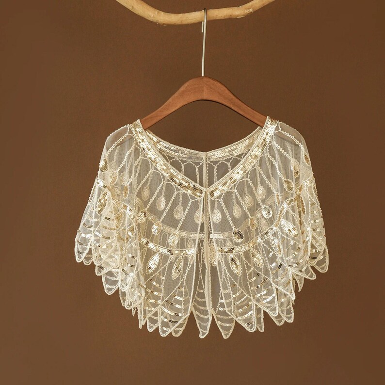 May include: A cream-colored, sequined shawl with a delicate, open-weave design. The shawl is embellished with gold sequins and hangs on a wooden hanger. The garment has a scalloped edge and a V-neck.