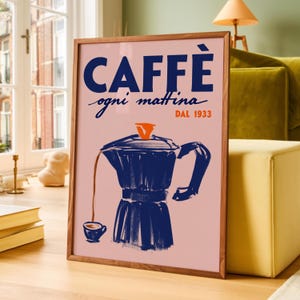 Op de afbeelding: Een ingelijste kunstprint met een lichtroze achtergrond en donkerblauwe tekst die CAFFÈ ogni mattina DAL 1933 leest. De illustratie toont een donkerblauwe mokkapot die koffie schenkt in een klein bijpassend kopje met een oranje deksel.