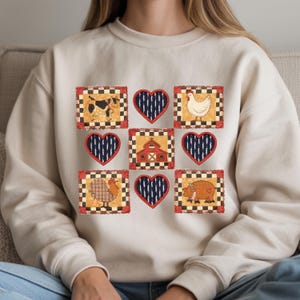 Könnte beinhalten: Cremefarbenes Sweatshirt mit einem Patchwork-Design aus Hoftieren und Herzen. Das Design umfasst eine Kuh, ein Huhn, einen Hahn, ein Schwein und eine Scheune, jeweils in einem karierten Rahmen. Die Herzen haben ein marineblaues Streifenmuster.