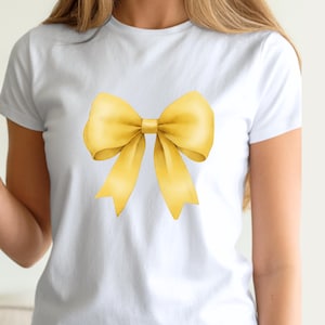 May include: White t-shirt featuring a large, watercolor-style yellow bow. The bow has detailed shading and highlights, giving it a realistic appearance. The shirt is a classic crew neck style, perfect for casual wear.