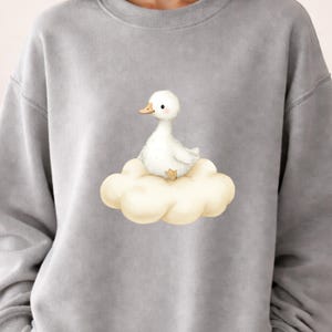 May include: A gray sweatshirt featuring a whimsical illustration of a white duckling perched on a fluffy cloud. The duckling has a yellow beak and feet. The design is centered on the chest.