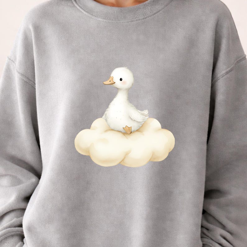 May include: A gray sweatshirt featuring a whimsical illustration of a white duckling perched on a fluffy cloud. The duckling has a yellow beak and feet. The design is centered on the chest.