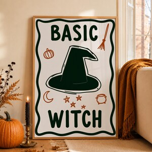 Può includere: Una stampa artistica da parete rettangolare in una cornice di legno chiaro con il testo BASIC WITCH in verde scuro. Il design include un cappello da strega verde scuro al centro, una piccola zucca arancione, una scopa, una luna crescente, stelle e un calderone.