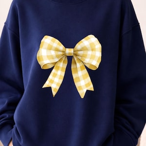 May include: Navy blue sweatshirt featuring a large yellow and white gingham bow design. The bow is centered on the front of the sweatshirt. The sweatshirt has a crew neck and long sleeves.